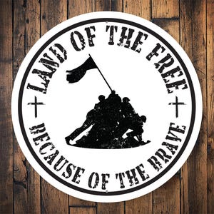 May include: A round, white sticker with a black graphic of soldiers raising a flag. The text "LAND OF THE FREE" and "BECAUSE OF THE BRAVE" are in a circular pattern around the image. The background is a wooden surface.