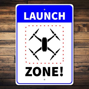 May include: A white sign with blue and red borders. The sign says "LAUNCH ZONE!" with a black drone icon inside a red dashed square.