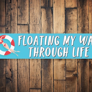 May include: A rectangular sign with the text "FLOATING MY WAY THROUGH LIFE" in white letters on a light blue background. The sign features an illustration of a person in a red swimsuit floating on a red and white inner tube. The background is a wooden surface.