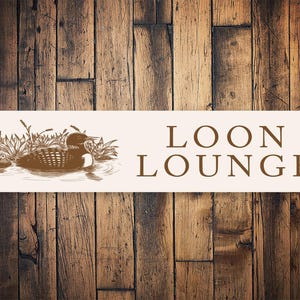 May include: A white sign with brown text and a brown illustration of a loon swimming in a lake. The sign reads "LOON LOUNGE".