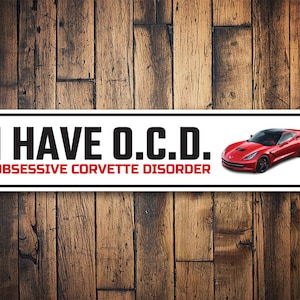 May include: White metal sign with the text "I HAVE O.C.D. OBSESSIVE CORVETTE DISORDER" and a red Corvette sports car on a wood background.
