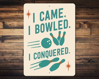 May include: Cream-coloured metal sign with the text "I CAME. I BOWLED. I CONQUERED." in teal. The sign features bowling pins, a bowling ball, and starburst designs. The sign is mounted on a wooden background.
