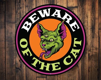 May include: A round, black and purple sign with the text "Beware of the Cat" in white and green lettering. A green, three-eyed cat with a purple ear and tongue is in the centre of the sign.