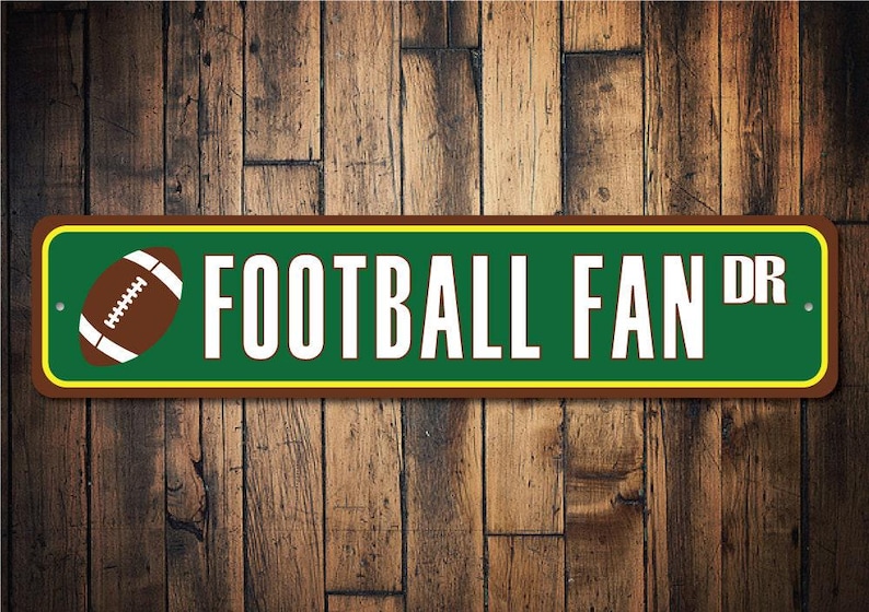May include: A green and brown metal sign with a yellow border. The sign reads "Football Fan Dr." and features a brown and white football graphic.