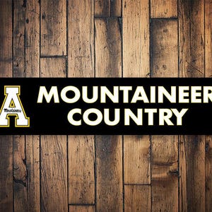 May include: Black and yellow sign with the text "MOUNTAINEER COUNTRY" and a yellow "A" with the word "Mountaineer" inside.