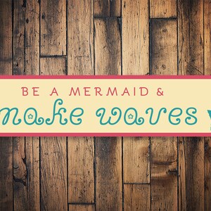 May include: A yellow and pink sign with the text "BE A MERMAID & make waves" in teal blue cursive font. A teal blue mermaid silhouette is on the right side of the sign.