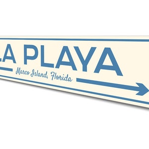 May include: A white and blue metal sign with the text "LA PLAYA" in blue letters. The sign also includes the text "Marco Island, Florida" in a smaller font. An arrow points to the right.
