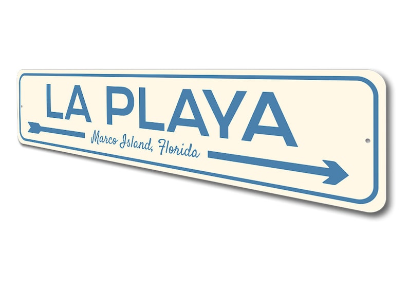 May include: A white and blue metal sign with the text "LA PLAYA" in blue letters. The sign also includes the text "Marco Island, Florida" in a smaller font. An arrow points to the right.