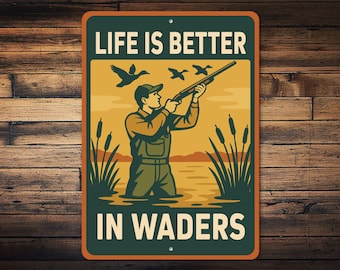 Op de afbeelding: Rechthoekig metalen bord met een vintage illustratie van een jager in waadpak die met een geweer op vliegende eenden richt. Het bord heeft de tekst "LIFE IS BETTER IN WADERS" in witte letters.