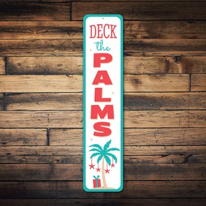 May include: A vertical metal sign with a turquoise border and white background. The sign says "Deck the Palms" in red letters with a palm tree with red ornaments and a red gift box at the base.