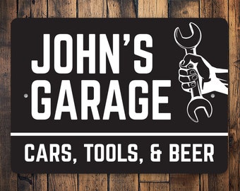 Puede incluir: Un letrero de metal negro y blanco con el texto "John's Garage Cars, Tools, & Beer". Una llave inglesa está representada en la esquina superior derecha del letrero.