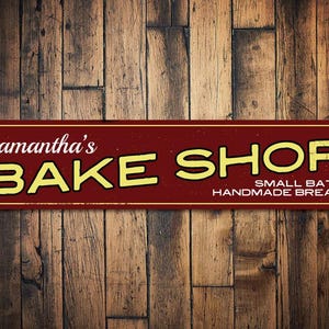 May include: A rectangular, maroon metal sign with the words "Samantha's BAKE SHOP" in white and yellow lettering. Below, it reads "SMALL BATCH HANDMADE BREADS". The sign is set against a rustic wooden background.