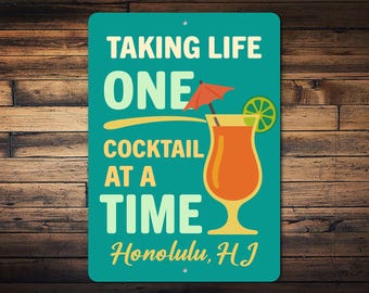 Op de afbeelding: Een turquoise metalen bord met de tekst "TAKING LIFE ONE COCKTAIL AT A TIME" en "Honolulu, HI". Het bord toont een cocktailillustratie met een oranje drankje, een schijfje limoen en een kleine paraplu.