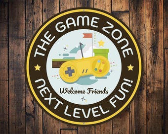 Peut inclure: Une pancarte ronde, marron et jaune avec le texte "The Game Zone Next Level Fun!" et une image de dessin animé d'une manette de jeu vidéo. La pancarte a un fond blanc avec un paysage de dessin animé et le texte "Welcome Friends".