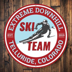 May include: Red and white circular metal sign with the text "Extreme Downhill Ski Team Telluride, Colorado Est. 2019" and a silhouette of a skier on a snowy mountain.