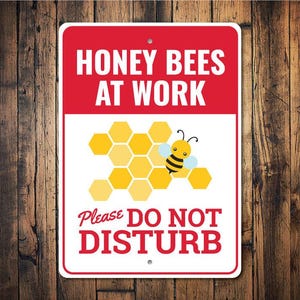 May include: A rectangular sign with a red top section that reads "HONEY BEES AT WORK" in white. Below is a white section with a yellow honeycomb and a cartoon bee. The bottom section reads "Please DO NOT DISTURB" in red.