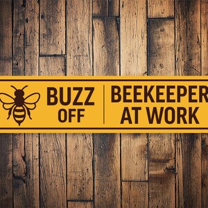 Può includere: Un cartello giallo con testo nero e un'immagine di un'ape. Il cartello dice "Buzz Off Beekeeper At Work".