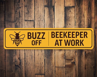 May include: Yellow sign with black text and a bee graphic. The sign reads "Buzz Off Beekeeper At Work".