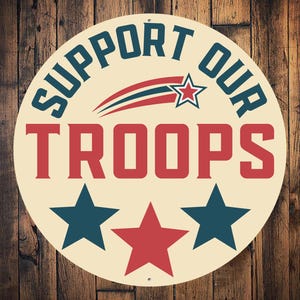 May include: A round, cream-colored sign with the text "Support Our Troops" in blue and red lettering. The sign also features a red, white, and blue star and a red, white, and blue shooting star. There are three stars at the bottom of the sign, two blue and one red.