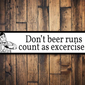 May include: A rectangular sign with the text "Don't beer runs count as excercise?" and a black and white illustration of a man pouring beer. The sign has a black border and is set against a wooden background.