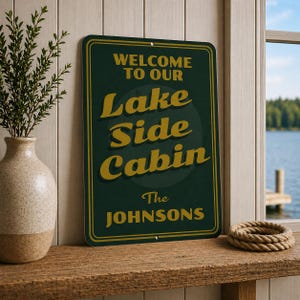 May include: A dark green metal sign with gold lettering that reads "WELCOME TO OUR LAKE SIDE CABIN The JOHNSONS". The sign is rectangular with rounded corners and a gold border. A decorative rope ring sits on a wooden shelf.