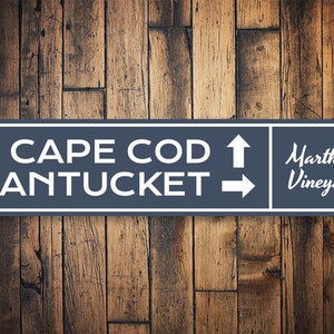 Peut inclure: Un panneau bleu et blanc avec du texte blanc et des flèches pointant dans différentes directions. Le panneau indique "CAPE COD", "NANTUCKET" et "Martha's Vineyard".