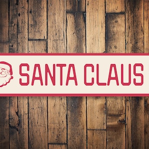 Op de afbeelding: Rood en wit metalen bord met een afbeelding van de kerstman en de tekst "SANTA CLAUS LN".