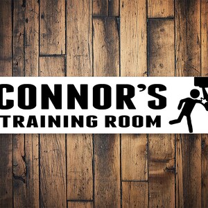 以下が含まれることがあります： 太字の黒い文字で「CONNOR'S TRAINING ROOM」と書かれた長方形の白い看板。ドアを突き破る人の白黒グラフィックが右側にあります。背景は木の壁です。