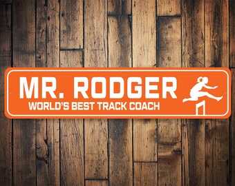 Puede incluir: Señal de metal naranja y blanca con el texto "MR. RODGER WORLD'S BEST TRACK COACH" y una silueta de una persona saltando sobre un obstáculo.