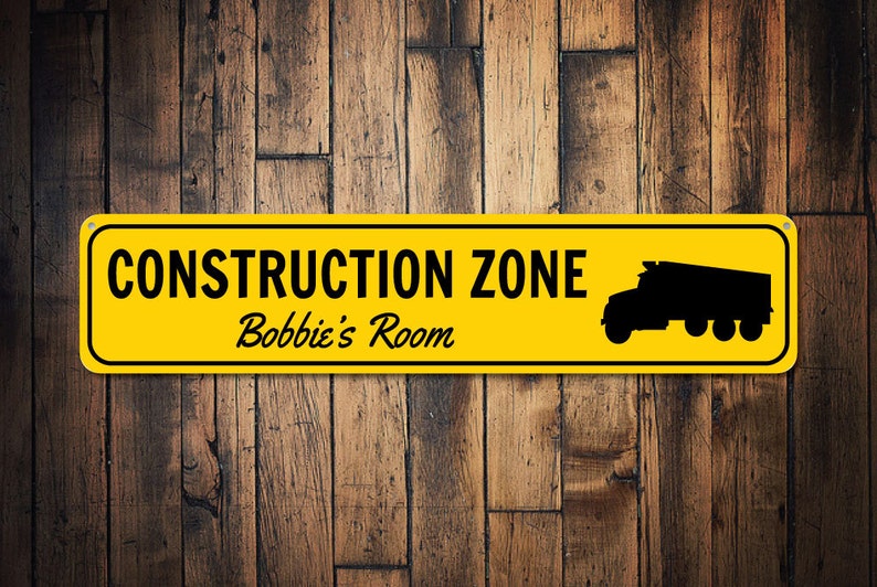 May include: A yellow construction zone sign with black text and a black silhouette of a dump truck. The sign reads "Construction Zone Bobbie's Room".