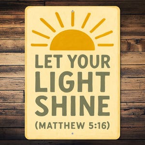 May include: Rectangular sign with a yellow background and a sun graphic. The text "LET YOUR LIGHT SHINE" is in green, with "(MATTHEW 5:16)" below. The sign is mounted on a wooden background.