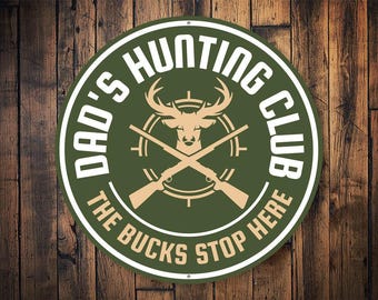 May include: A green and white round metal sign with the text "Dad's Hunting Club The Bucks Stop Here" and a silhouette of a deer with crossed rifles.