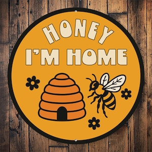 May include: A round, yellow sign with black trim and the text "Honey I'm Home" in white. The sign also features a beehive, a bee, and flowers.