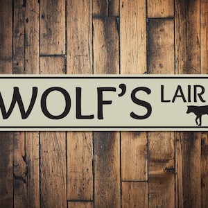 May include: A beige metal sign with black text and a black silhouette of a wolf. The sign reads "Wolf's Lair".