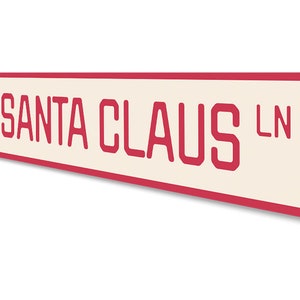 Op de afbeelding: Een rood en wit metalen bord met afgeronde hoeken waarop "SANTA CLAUS LN" staat met een cartoonafbeelding van de kerstman in de linkerbovenhoek.