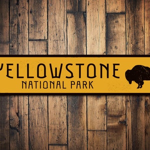 May include: Yellowstone National Park sign featuring a black silhouette of a bison against a yellow background. The sign is made of metal and has a distressed appearance.