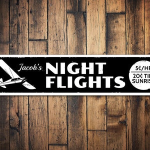 Puede incluir: Un letrero en blanco y negro con el texto "Jacob's Night Flights" y una pequeña ilustración de un avión. El letrero también incluye el texto "5¢/HR 20¢ TILL SUNRISE".