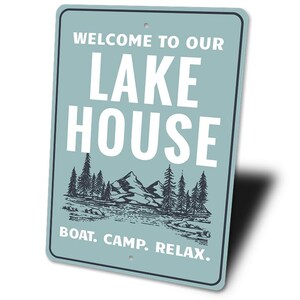 May include: A rectangular sign with rounded corners, featuring the text "WELCOME TO OUR LAKE HOUSE" in white block letters. Below, a graphic of mountains and trees is displayed. The bottom reads "BOAT. CAMP. RELAX."