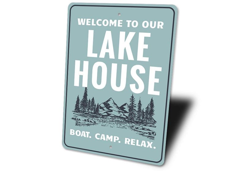 May include: A rectangular sign with rounded corners, featuring the text "WELCOME TO OUR LAKE HOUSE" in white block letters. Below, a graphic of mountains and trees is displayed. The bottom reads "BOAT. CAMP. RELAX."