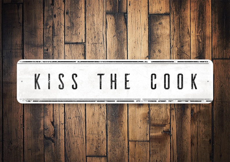 May include: White sign with black lettering on a wood background. The sign reads "Kiss the Cook."