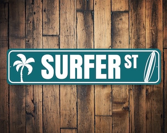 Puede incluir: Un letrero de calle turquesa con las palabras "SURFER ST" en blanco, junto con una palmera blanca y un gráfico de tabla de surf. El letrero está sobre un fondo de madera rústica.