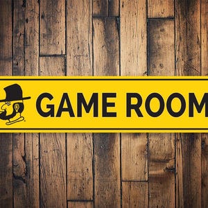 以下が含まれることがあります： 「GAME ROOM」の文字が黒で書かれた長方形の黄色い看板。帽子とパイプを持った男性の漫画の画像が特徴です。背景は木製の壁です。