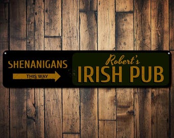 Puede incluir: Un letrero de metal negro y verde con letras doradas. El letrero dice "Shenanigans This Way" con una flecha apuntando hacia la derecha y "Robert's Irish Pub".