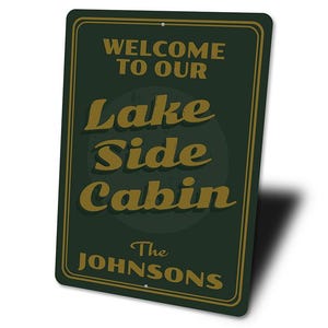 May include: A rectangular dark green metal sign with a gold border. The sign reads "WELCOME TO OUR LAKE SIDE CABIN" in gold lettering, with "The Johnsons" below. The sign has two holes for hanging.
