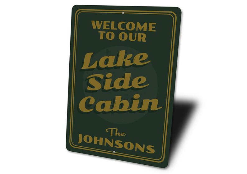 May include: A rectangular dark green metal sign with a gold border. The sign reads "WELCOME TO OUR LAKE SIDE CABIN" in gold lettering, with "The Johnsons" below. The sign has two holes for hanging.