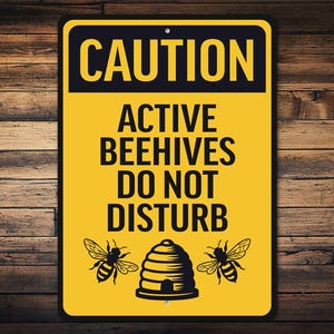May include: A rectangular yellow sign with black text that reads "CAUTION ACTIVE BEEHIVES DO NOT DISTURB." The sign features illustrations of two bees and a beehive. The sign is mounted on a wooden background.