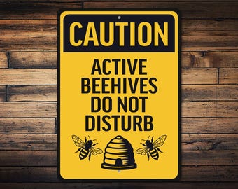 May include: A rectangular yellow sign with black text that reads "CAUTION ACTIVE BEEHIVES DO NOT DISTURB." The sign features illustrations of two bees and a beehive. The sign is mounted on a wooden background.