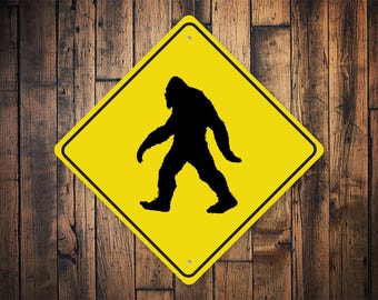 Puede incluir: Señal de advertencia amarilla en forma de diamante con una silueta negra de Bigfoot caminando. La señal es de metal y tiene un aspecto desgastado.