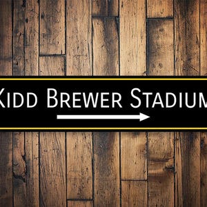 May include: Black and yellow sign with white text and an arrow pointing to the right. The sign reads "Kidd Brewer Stadium."
