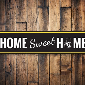 以下が含まれることがあります： 黒と黄色の金属製の看板に、白い文字で「HOME Sweet HOME」と書かれています。テキストの右側に帽子をかぶった漫画のキャラクターがいます。
